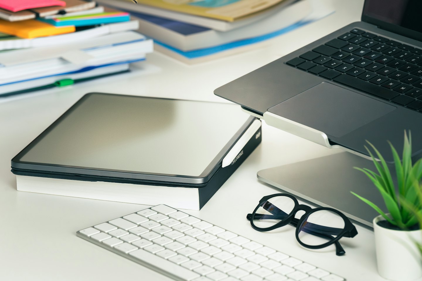 Laptop, tablet, keyboard, and glasses on a white desk.